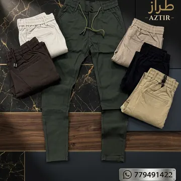 Six folded men's drawstring pants in various earth tones displayed on marble and wood surfaces