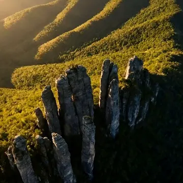 An aerial view of a forested mountain range with prominent rock spires illuminated by sunrise or sunset light