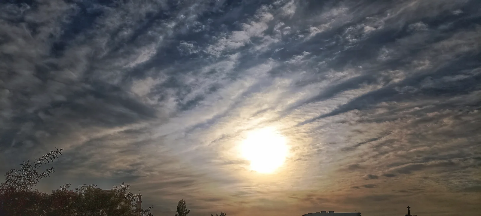 Sunlight breaks through textured clouds over urban skyline and trees at dawn or dusk