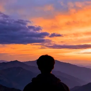 A person in silhouette stands on a mountain ridge facing a colorful sunset sky with layered mountain ranges in the background