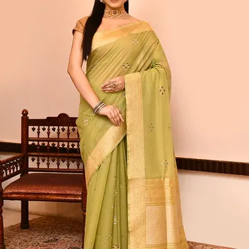 Woman wearing green embroidered saree with gold border standing beside wooden chair on patterned rug