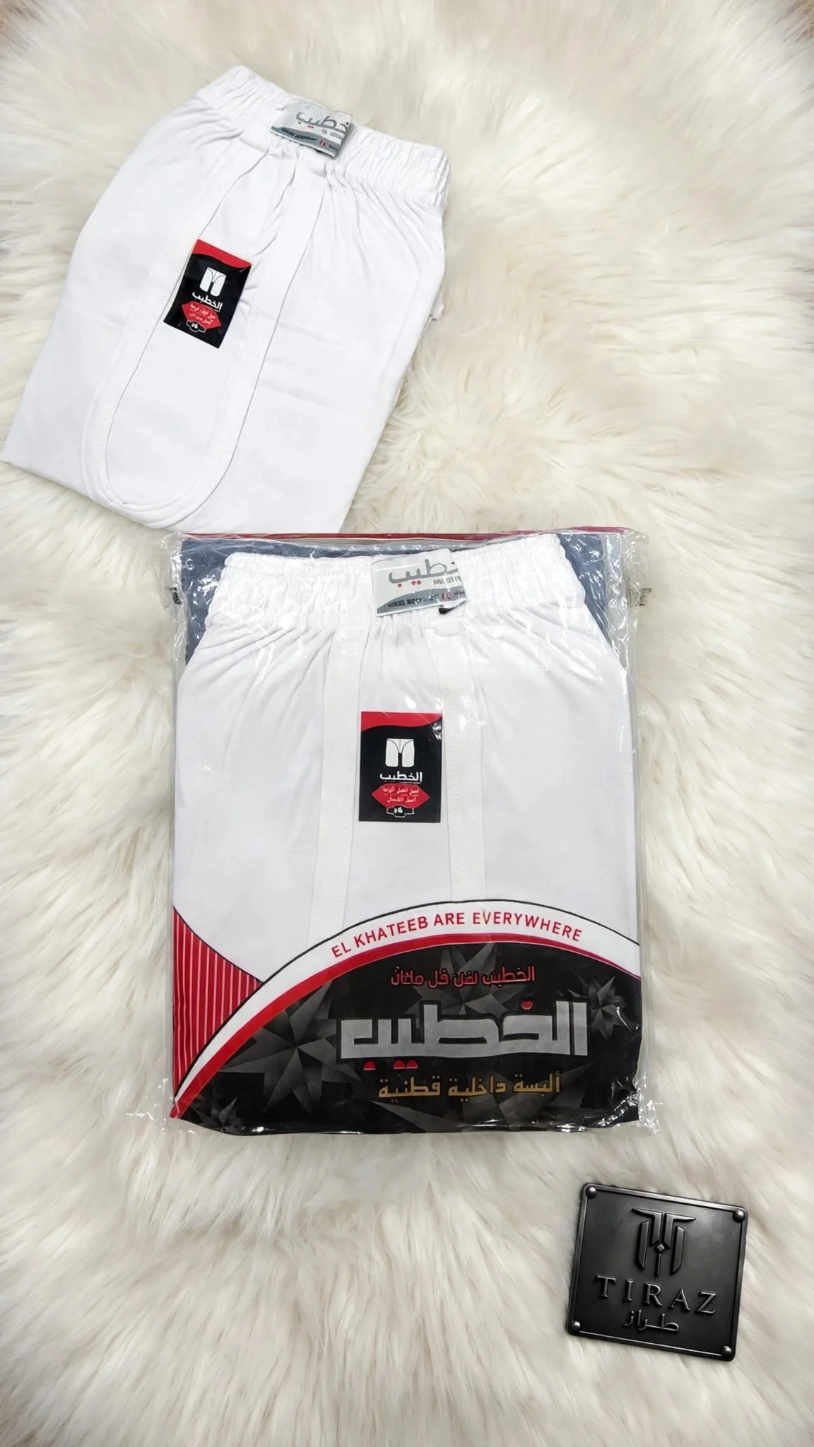 Two white El Khateeb shorts one folded one packaged on fluffy white rug with TIRAZ plaque
