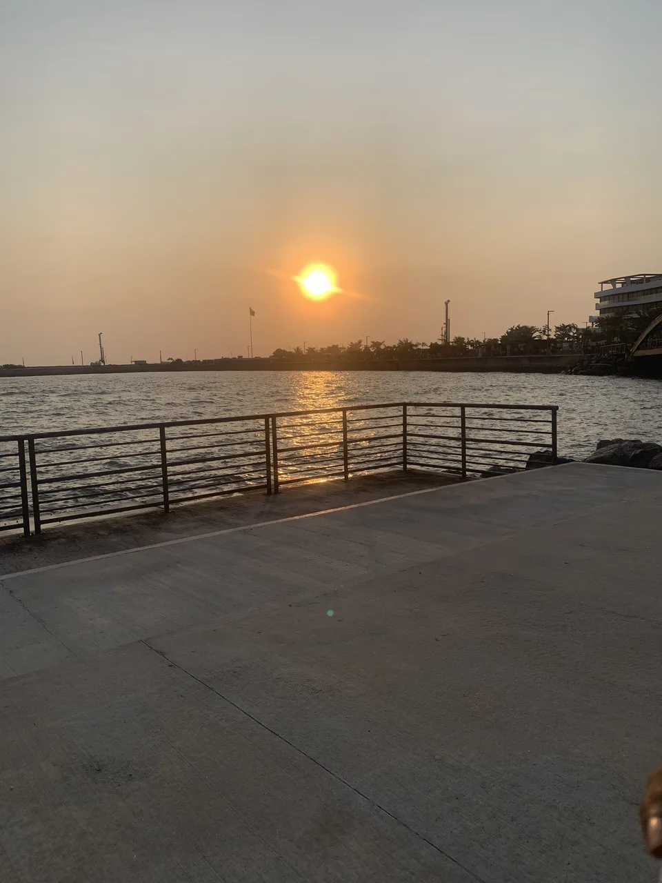 A sunset over a body of water viewed from a concrete pier with a metal railing, with buildings and industrial structures visible in the distance