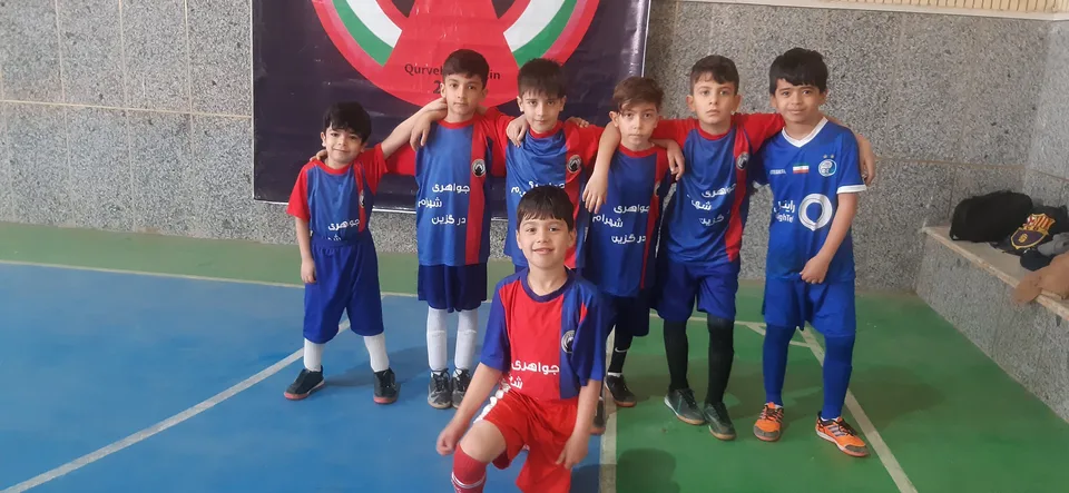A group of young boys in blue and red soccer uniforms pose for a photo indoors on a court, with a banner behind them