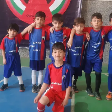 A group of young boys in blue and red soccer uniforms pose for a photo indoors on a court, with a banner behind them