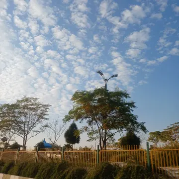 A low-angle view of a blue sky with scattered white clouds, framed by trees, a yellow fence, and a streetlamp