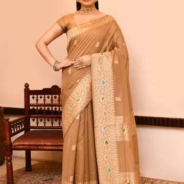 Woman wearing beige embroidered saree with gold jewelry standing beside wooden chair on patterned rug