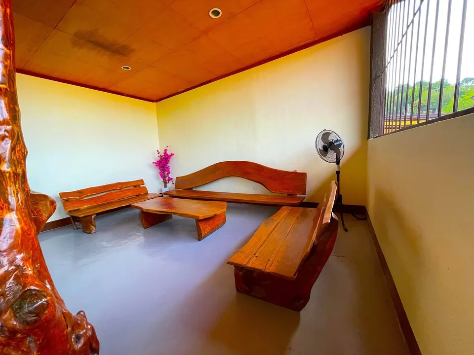 A room with wooden benches and table, a standing fan, and a pink flower vase near a window