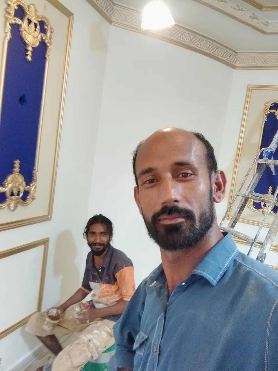 Two men in a room with ornate gold-trimmed blue wall panels and a ladder