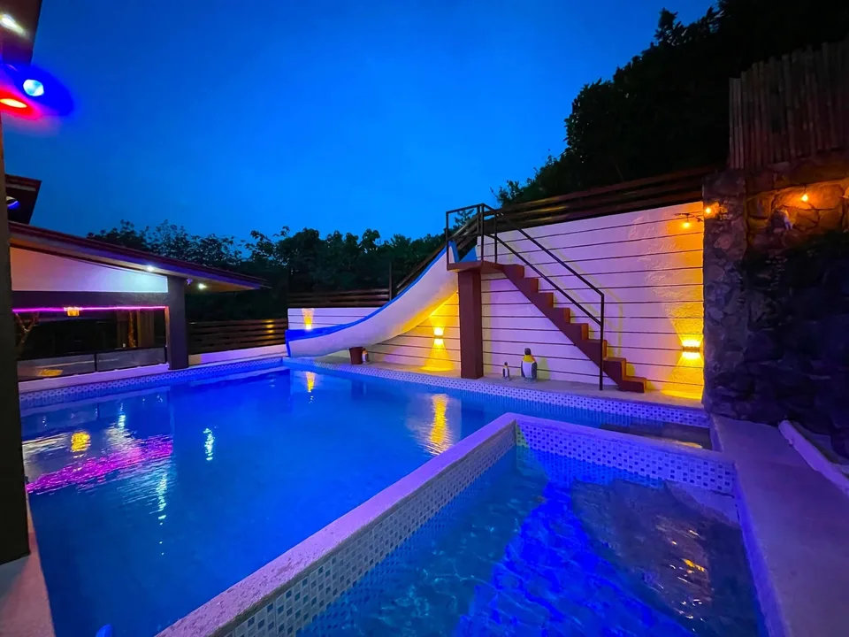 A brightly lit pool with a slide and stairs at night surrounded by trees and stone walls