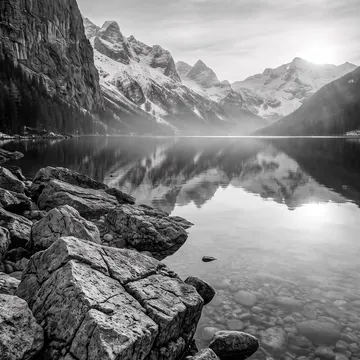 Serene black and white mountain lake with rocky foreground and snowcapped peaks reflected in calm water