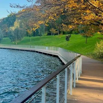 Curved wooden boardwalk with white railings along a lake with autumn trees and blue sky