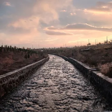 A low-angle view of a concrete-lined water channel flowing through a dry, vegetated landscape under a cloudy sunset sky